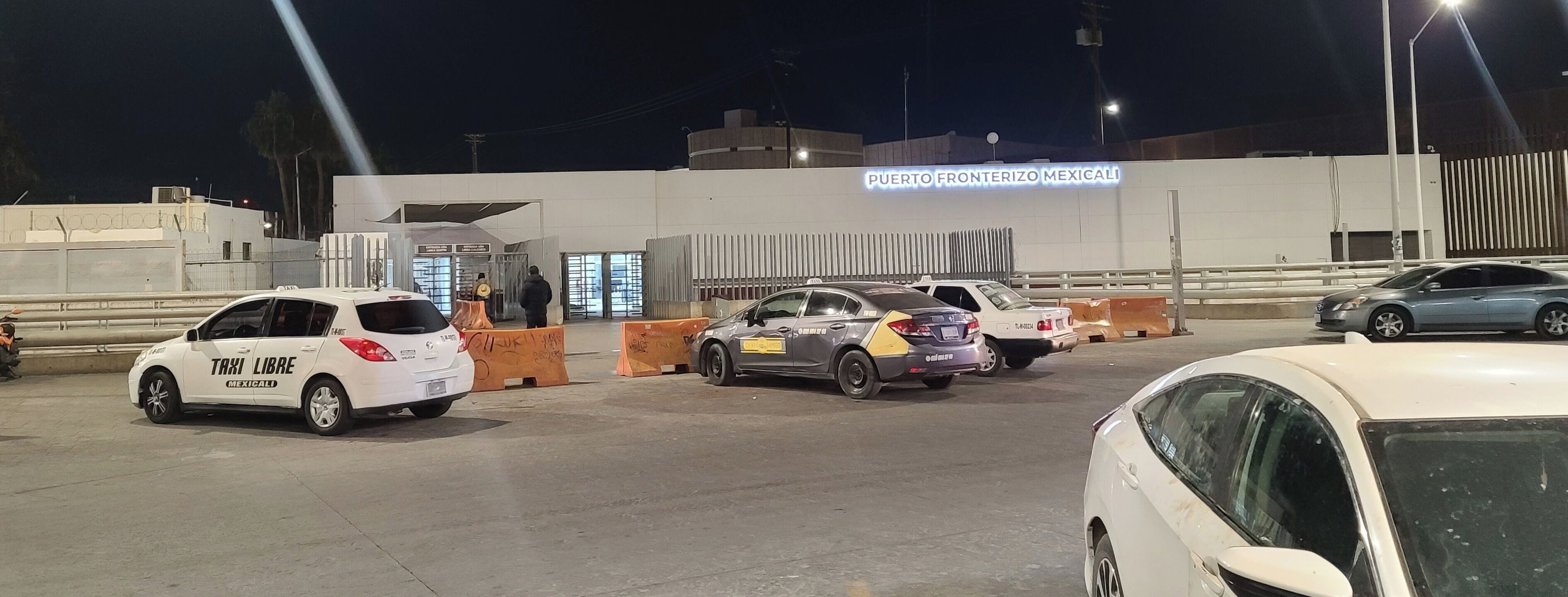 A nighttime scene at a toll booth with multiple cars and a sign reading "Puerto Fronterizo Mexicali".