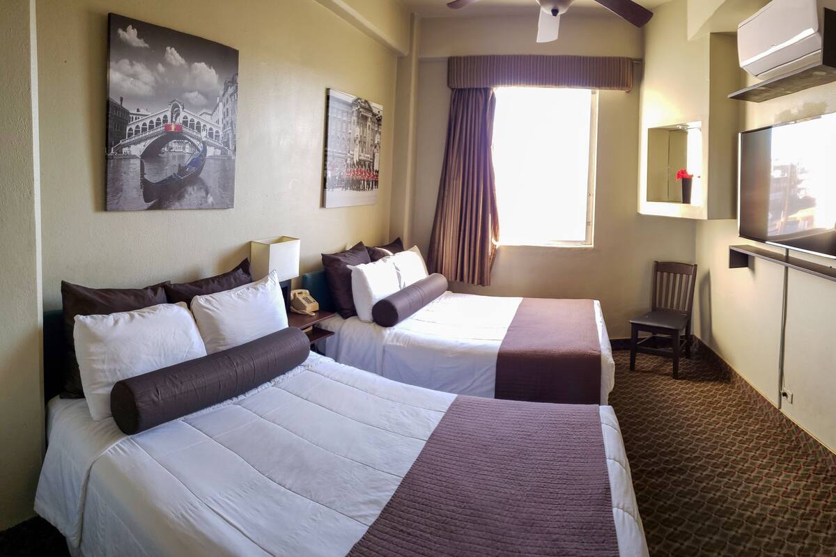 A hotel room with two beds, a TV, a bathroom with a shower, and a window with curtains.