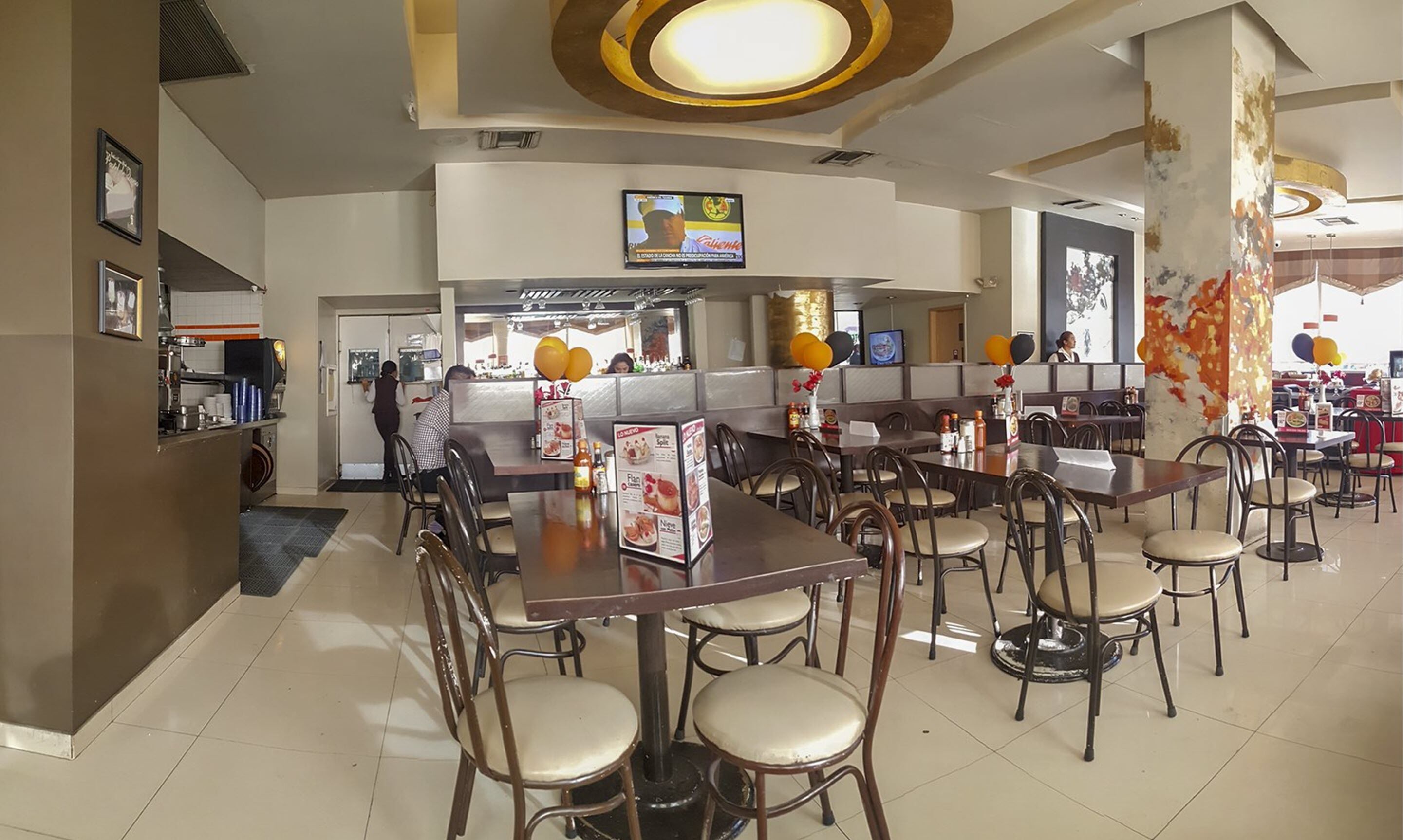Ein Restaurant mit mehreren Tischen, Stühlen, einer Theke mit Speisekarte und einem Fernsehbildschirm.