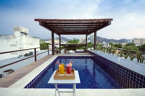 Piscina no terraço com pergolado de madeira, mesa com duas bebidas e vista para a cidade.