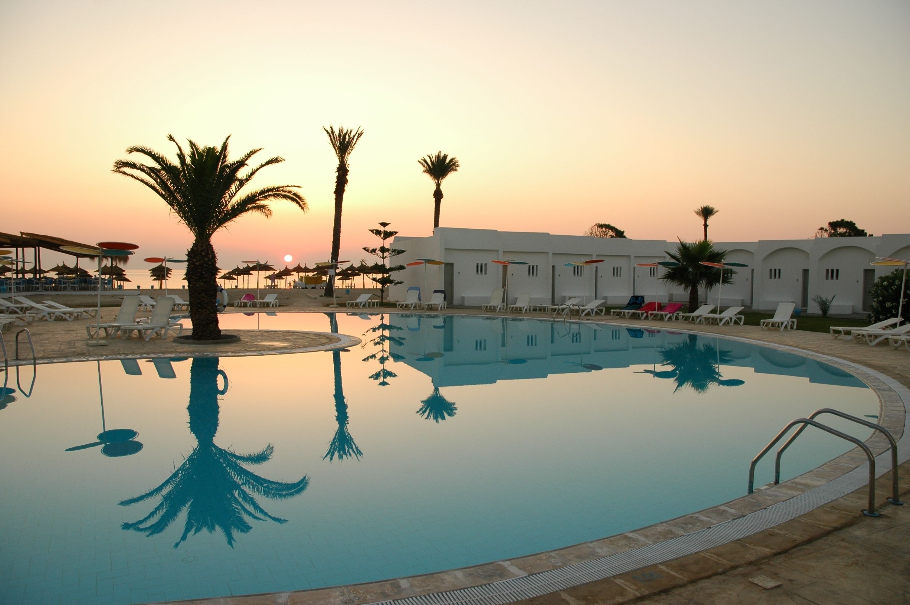 A swimming pool with lounge chairs and umbrellas, surrounded by palm trees and a building with a white facade.