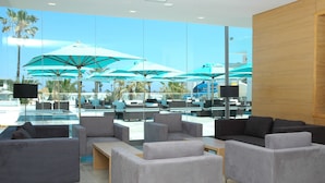 A modern lounge area with grey sofas and tables, large windows with a view of a pool and umbrellas. - Thalassa Sousse Resort & Aquapark (Sousse)