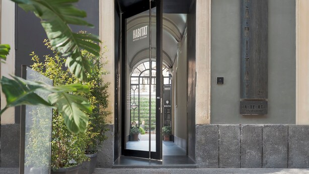 L'ingresso di un hotel con una porta in vetro, un cartello con su scritto "HABITAT MATERIA SICILANA" e una pianta in vaso a sinistra.