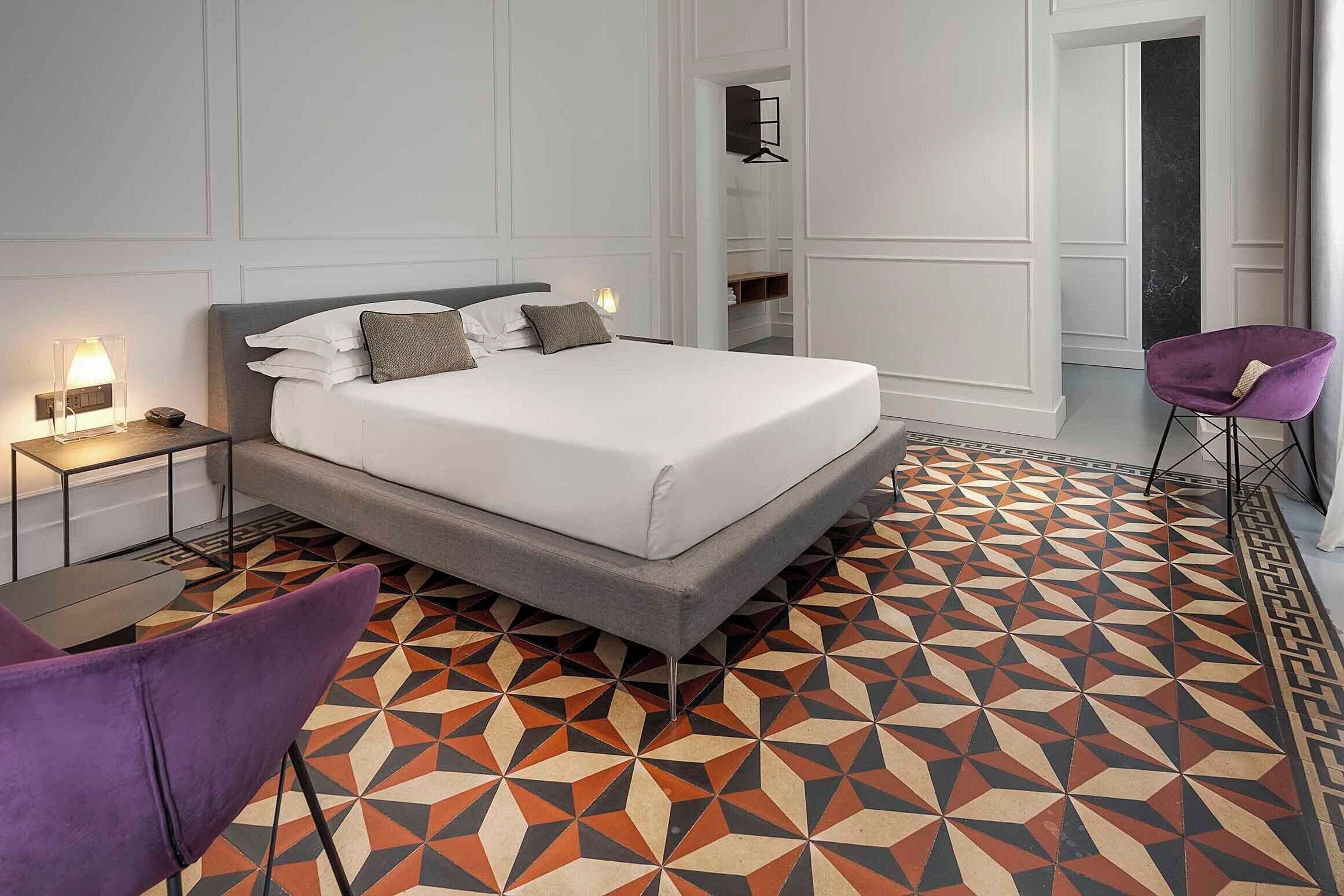 A modern hotel room with a large bed, a purple chair, and a patterned floor.