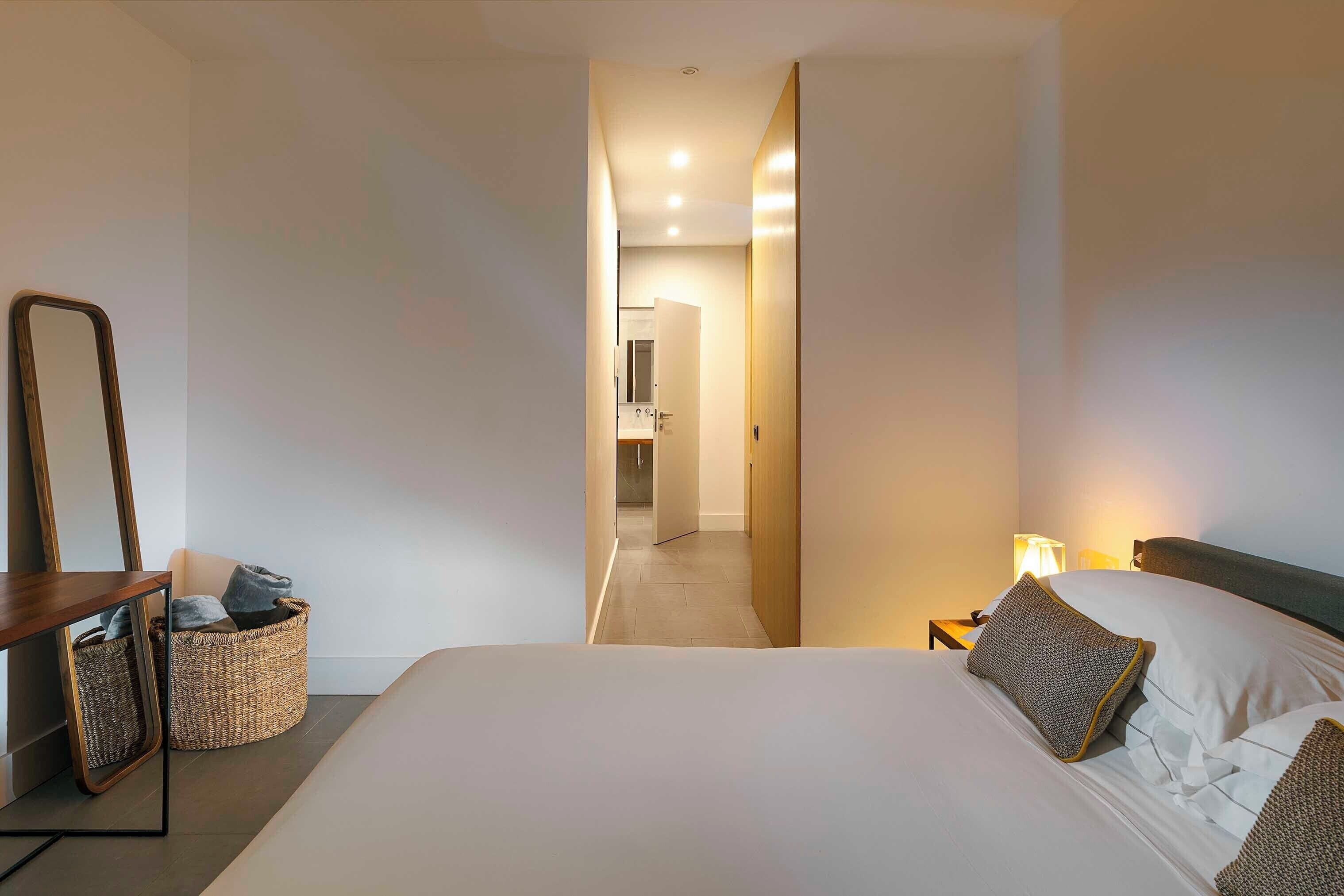 Habitación de hotel con una cama grande, una mesita de noche con lámpara, un espejo y una cesta de mimbre.