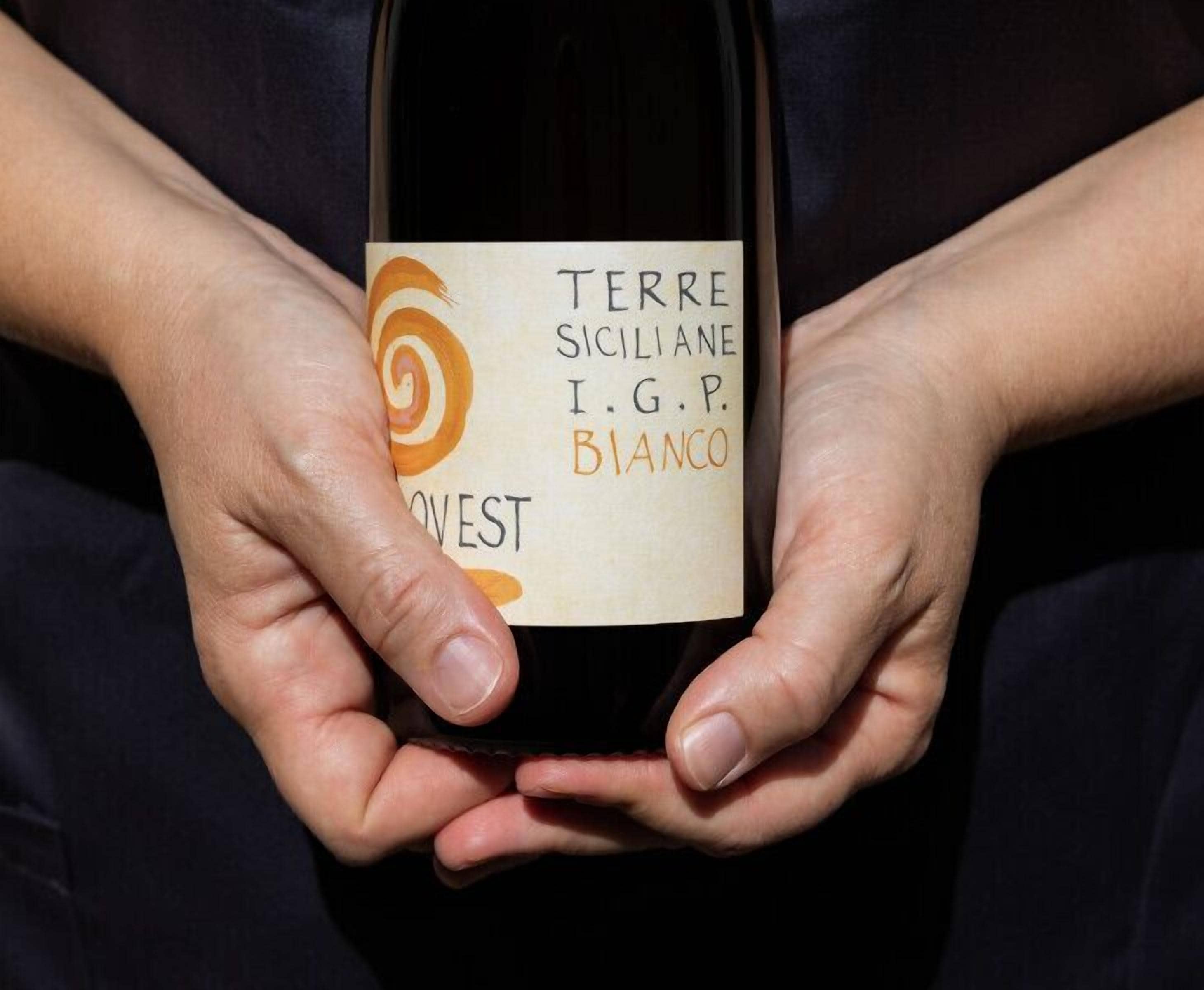 Una persona che tiene in mano una bottiglia di vino con un'etichetta che recita “TERRE SICILIANE I.G.P. BIANCO”.
