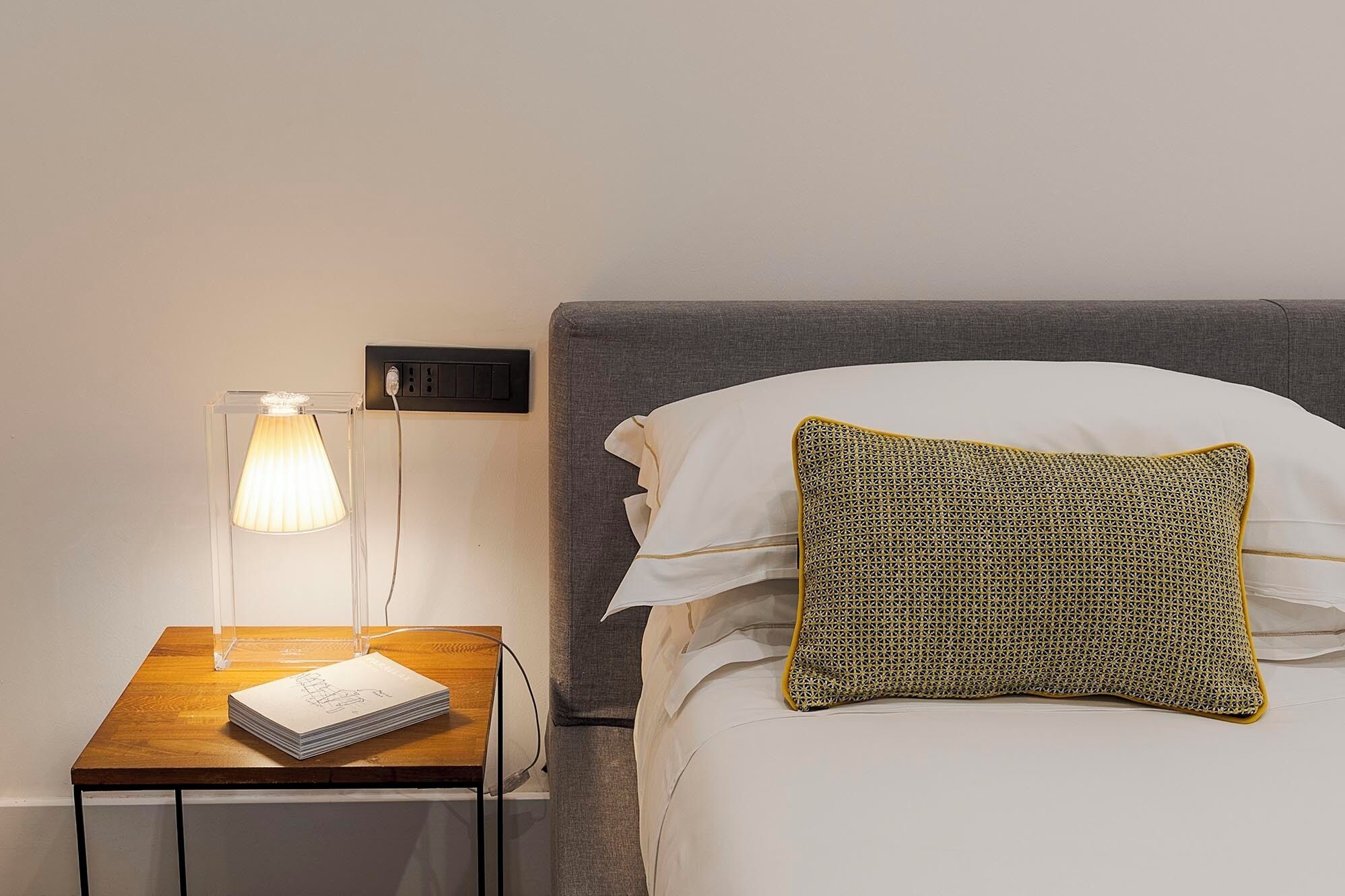 A neatly made bed with a textured pillow, a bedside lamp, and a stack of papers on a wooden nightstand.