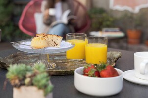 Un desayuno con una rebanada de pastel en un plato de vidrio, dos vasos de jugo de naranja, un bol de fresas y una taza blanca sobre una mesa de madera rústica.