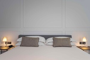 Una cama bien hecha con sábanas blancas, dos almohadas estampadas y un cabecero gris. Hay dos mesitas de noche con lámparas a cada lado de la cama.