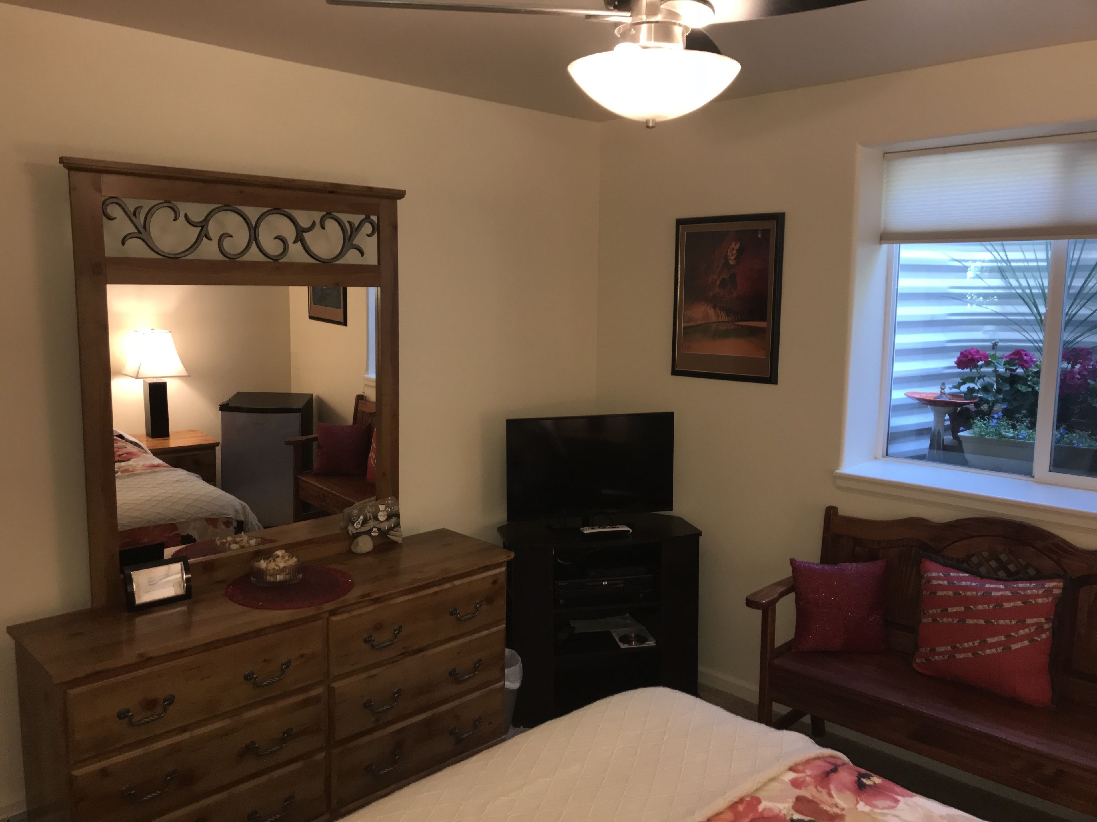 Een slaapkamer met een houten dressoir, een televisie, een bed met een bloemenprint dekbed, een raam met uitzicht op de omgeving en een plafondventilator.