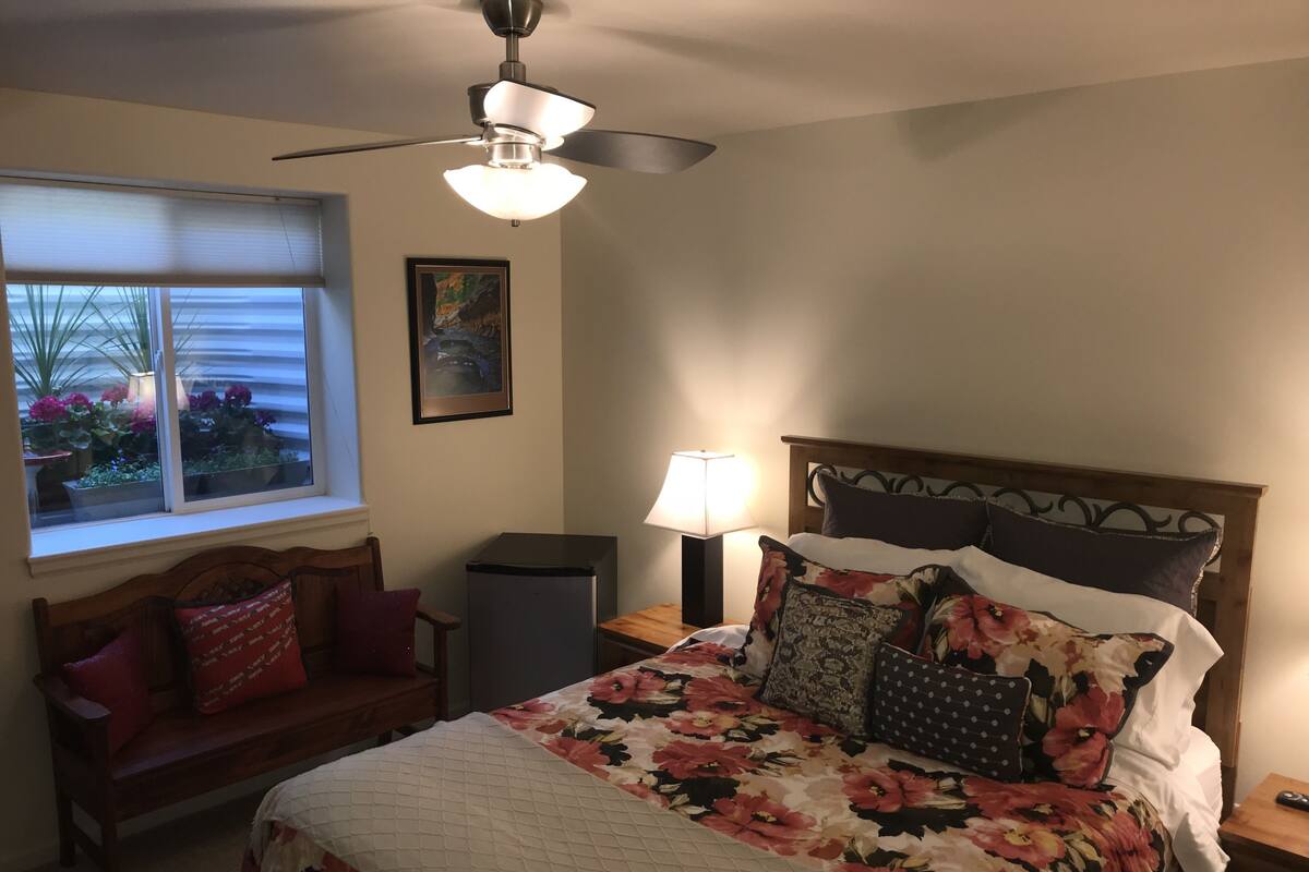 A bedroom with a bed, a nightstand, a lamp, a ceiling fan, a window with blinds, a framed picture, and a bench.