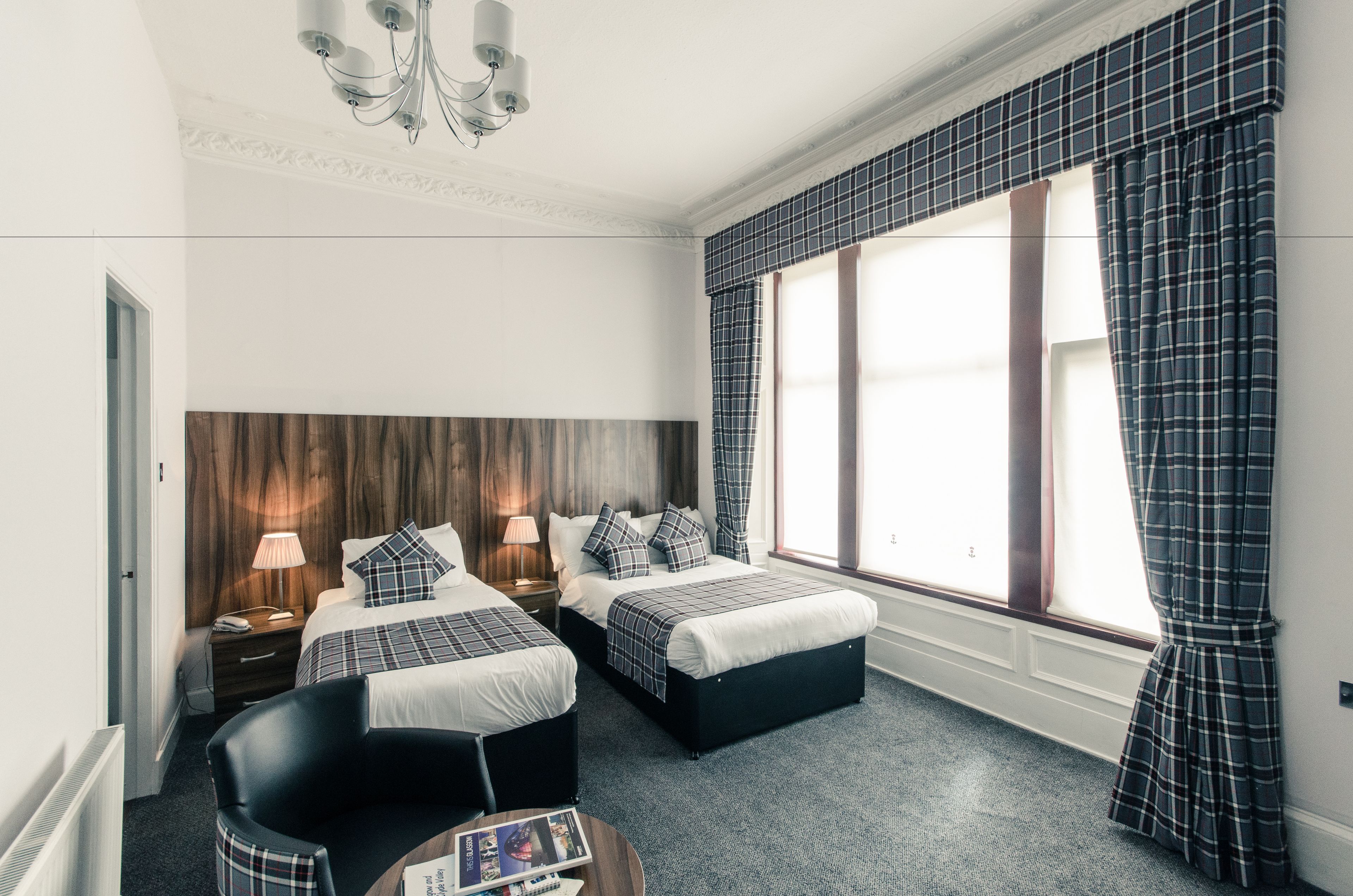 A hotel room with two beds, a wooden headboard, a chandelier, a large window with curtains, and a small table with a magazine.