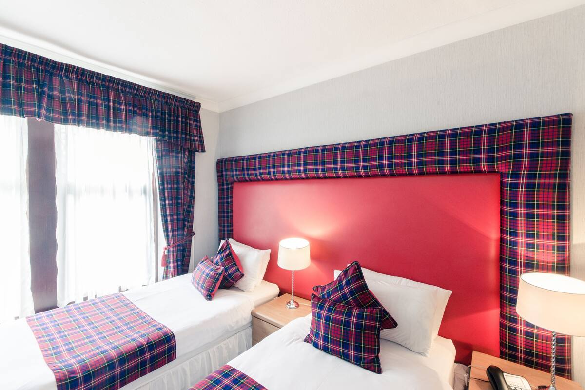 A hotel room with two beds, red headboard, plaid curtains, and a bedside lamp.