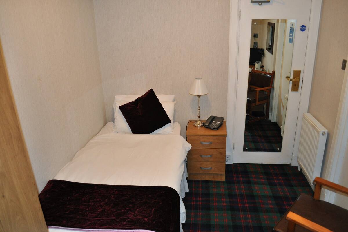 A small, neatly arranged bedroom with a bed, bedside table, lamp, and a mirror.