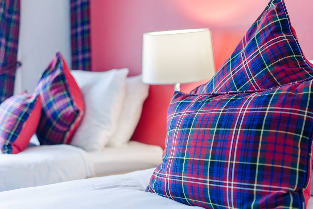 A bed with white bedding and multiple plaid pillows in a room with a red wall and a white lamp.