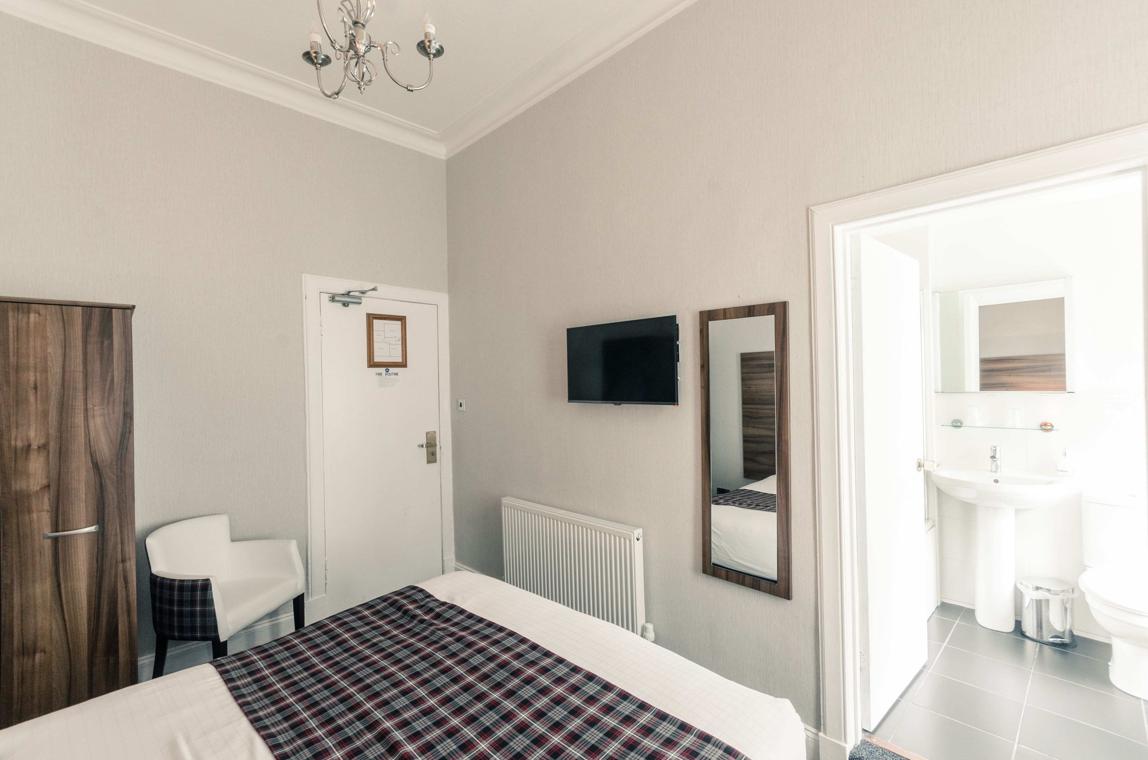 Une chambre d’hôtel avec un lit, une chaise, une télévision, un miroir et une salle de bain visible à travers une porte ouverte.