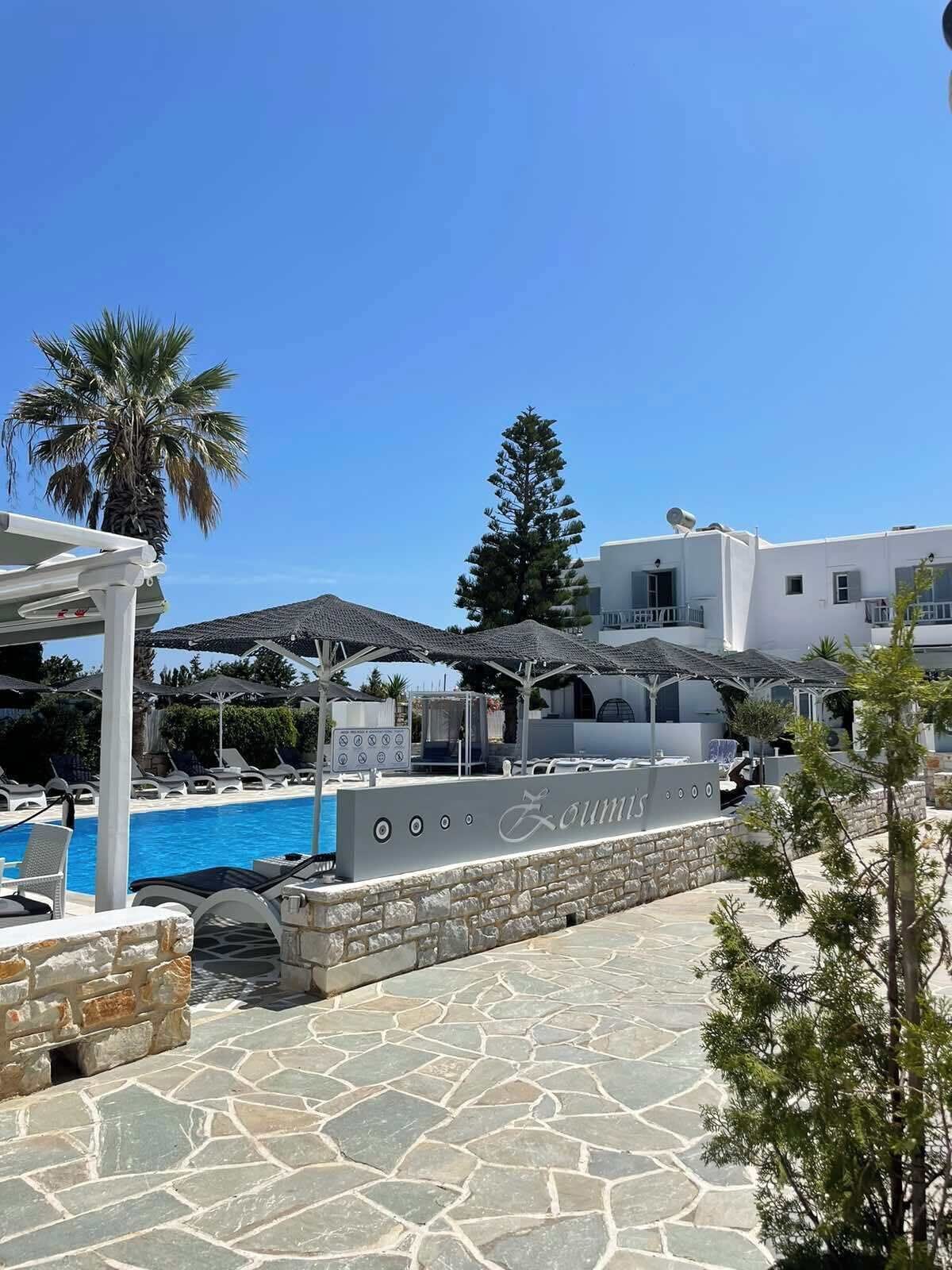 A pool area with lounge chairs and umbrellas, a stone wall with the name 'Zoumis', and a building in the background.