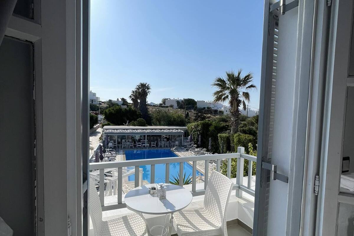 A balcony with a white table and chairs, overlooking a pool area with lounge chairs and a clear blue sky.