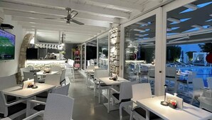 An indoor dining area with white tables and chairs, a ceiling fan, and a view of a pool through large windows.