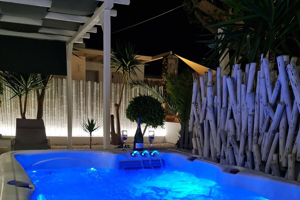 A hot tub with blue lighting, surrounded by a white pergola, palm trees, and a bamboo fence.