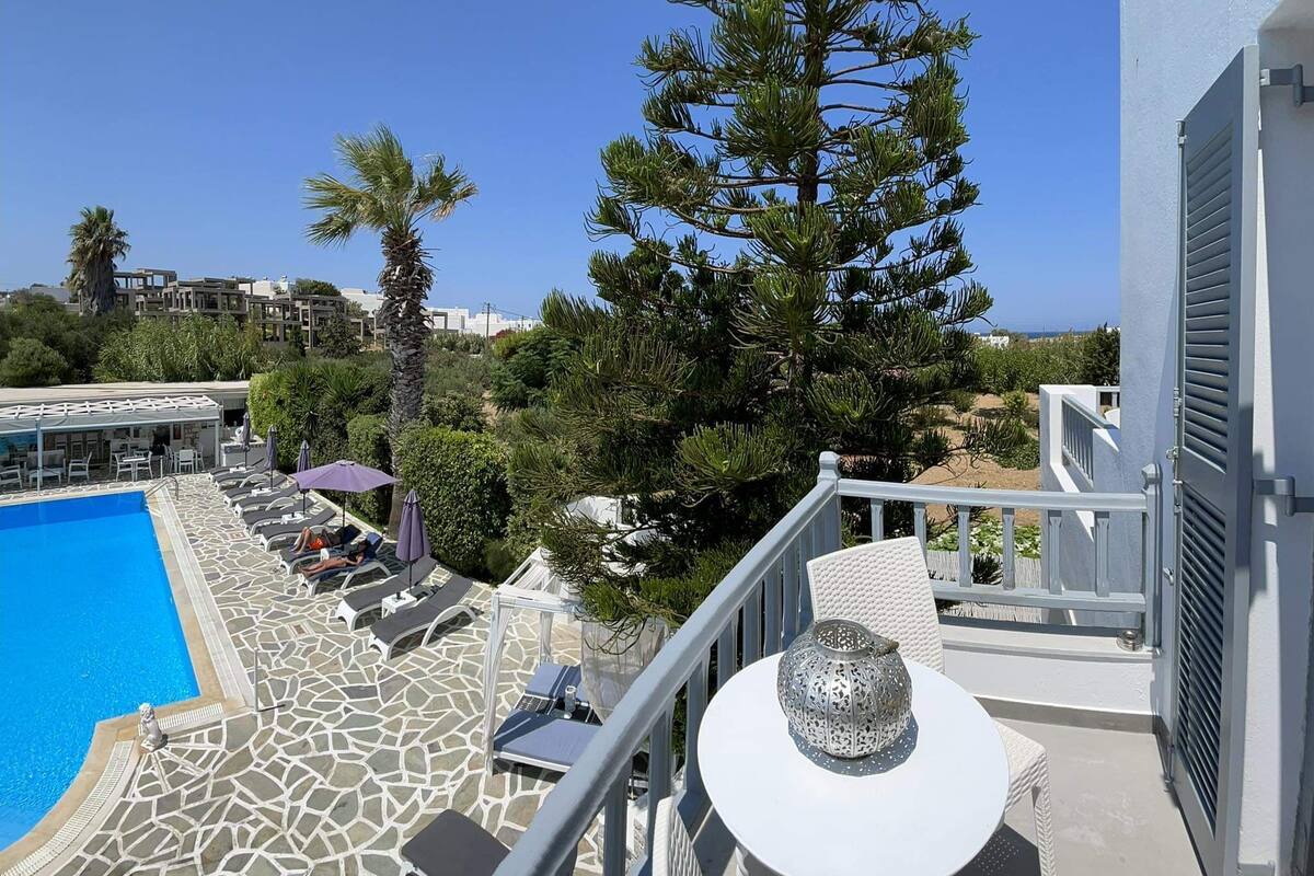 A poolside area with lounge chairs and a table, a large tree, and a view of the pool and surrounding buildings.
