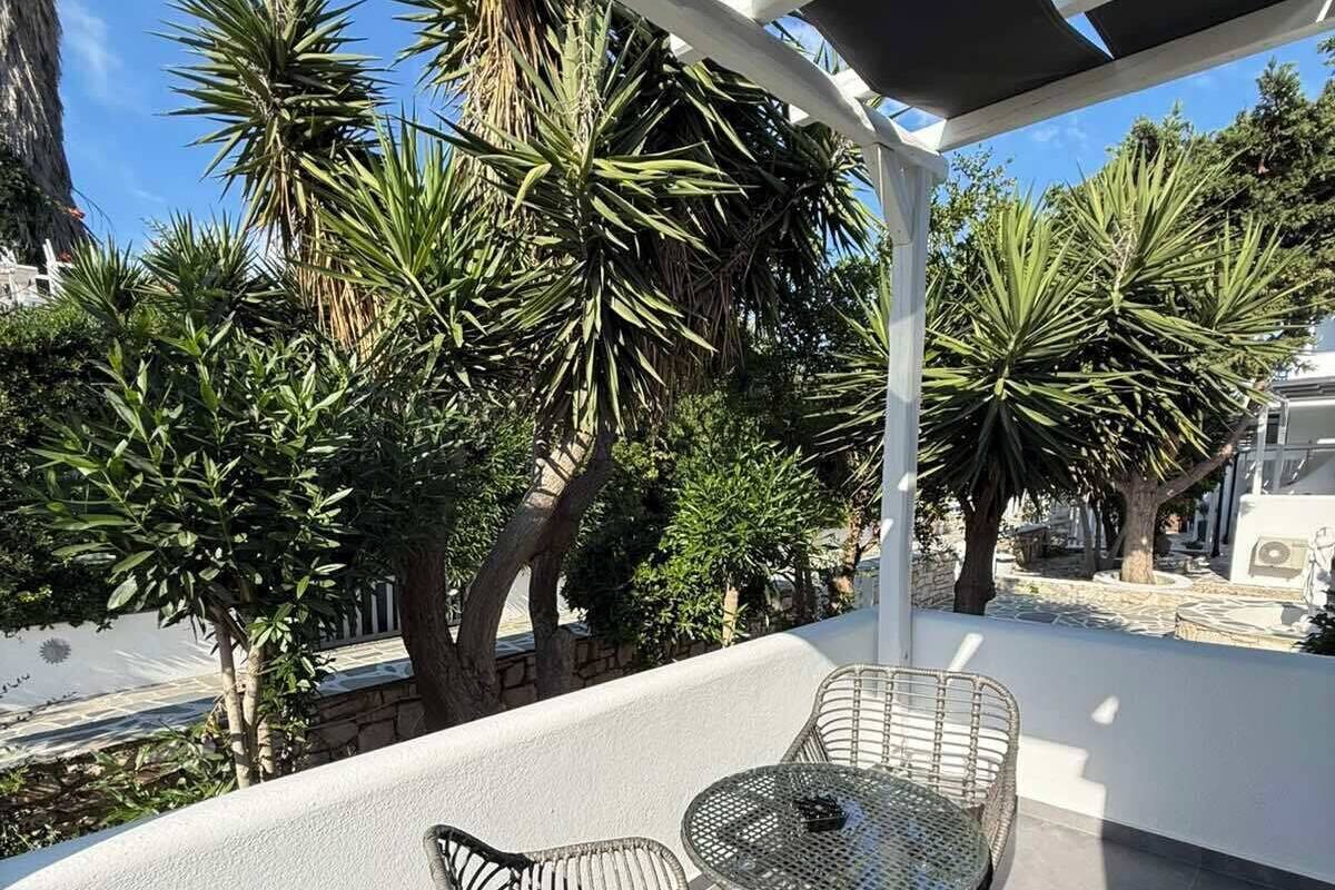 An outdoor patio with a table and two wicker chairs, surrounded by tropical plants and a white pergola.