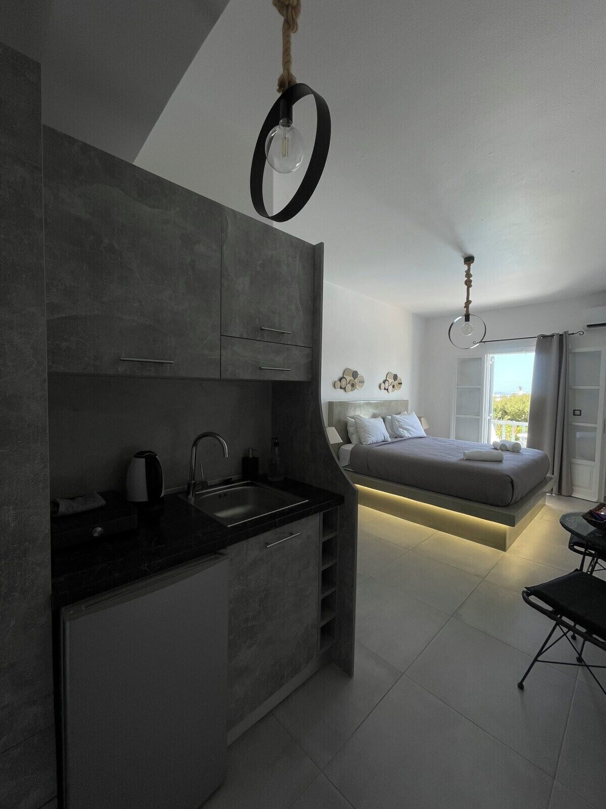 A modern kitchen with dark cabinets and a black countertop, a bed with a gray mattress and white pillows, and a dining area with a table and chairs.