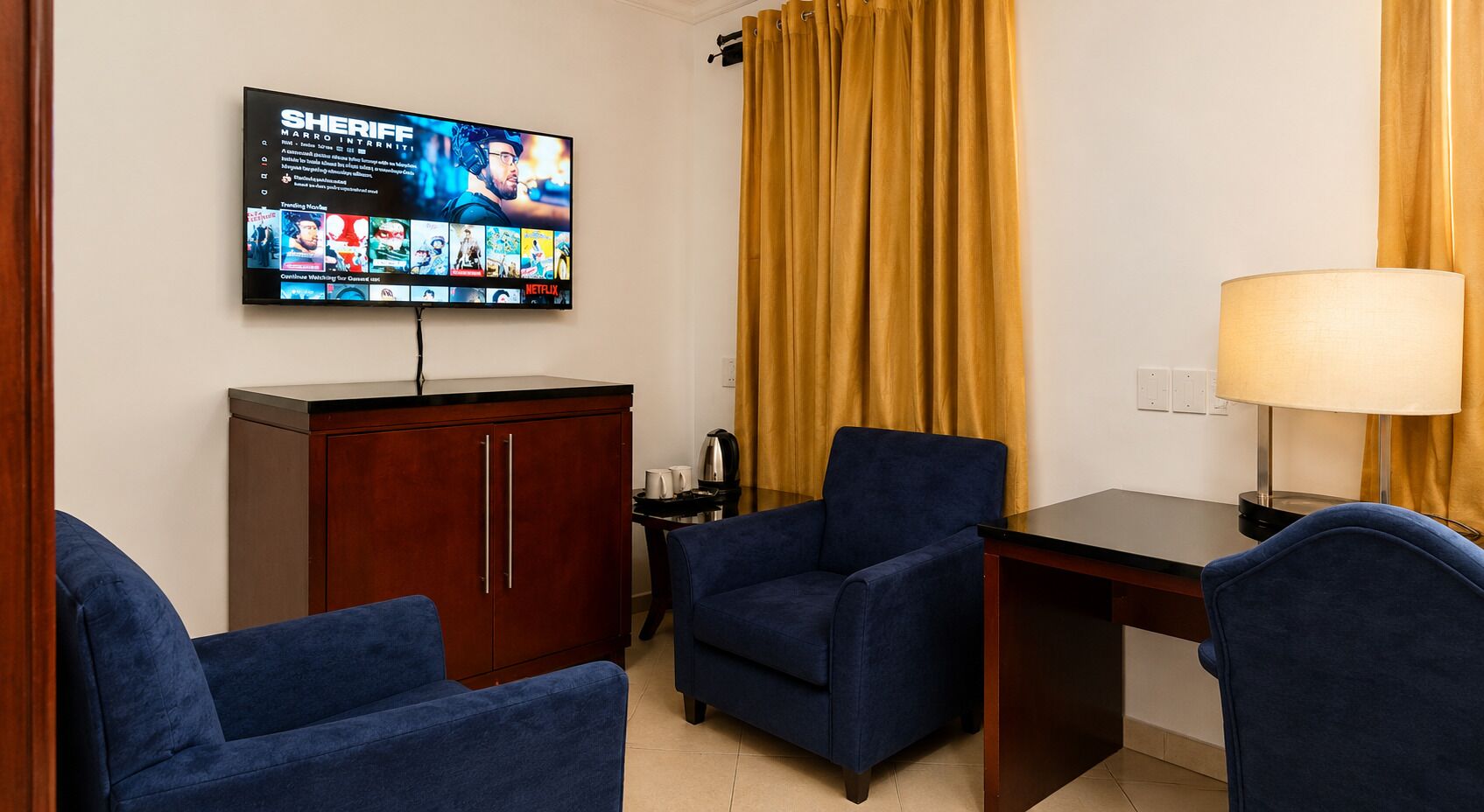 A hotel room with a flat-screen TV displaying a streaming service interface, a wooden cabinet, two blue armchairs, a small table, and a lamp.