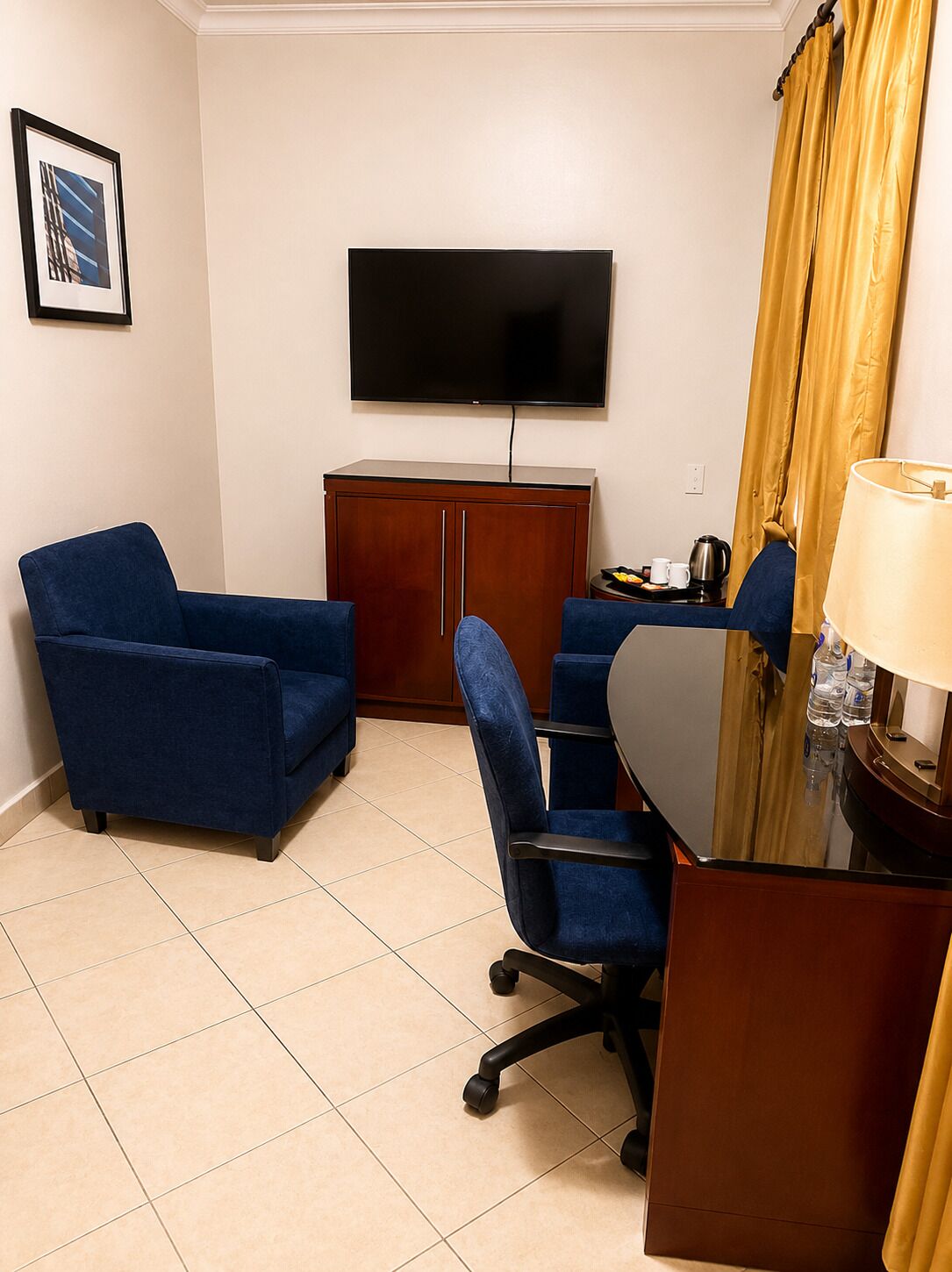 Une chambre d’hôtel équipée d’un téléviseur à écran plat fixé au mur, de deux fauteuils bleus, d’une armoire en bois, d’une petite table avec une bouilloire et des tasses, et d’une lampe.