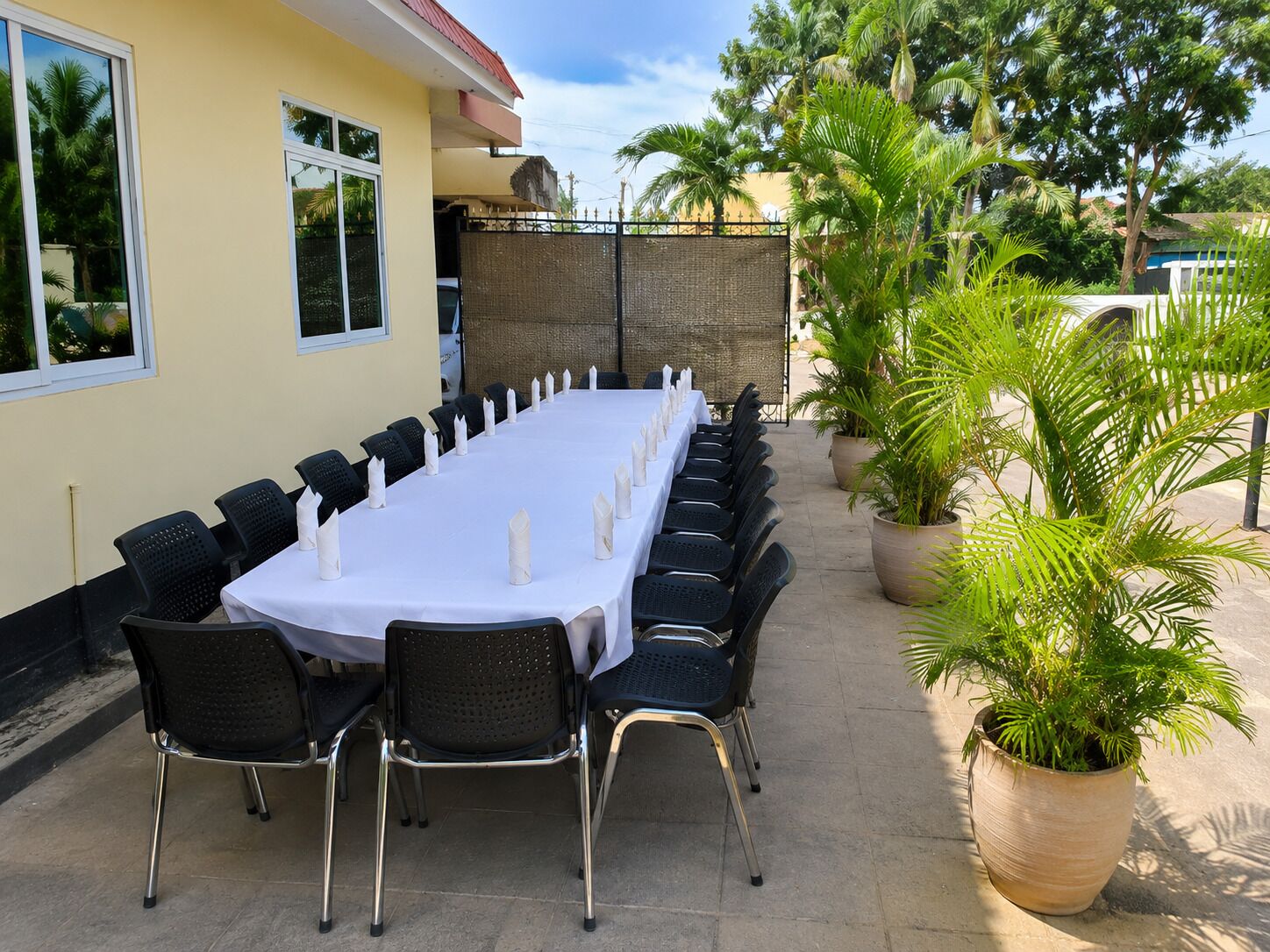 A long dining table set for a meal with chairs, a building with a red roof, and potted plants.