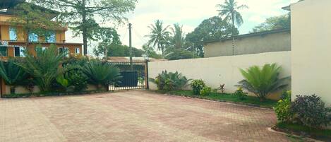A paved courtyard with a gated entrance, surrounded by palm trees and various plants.