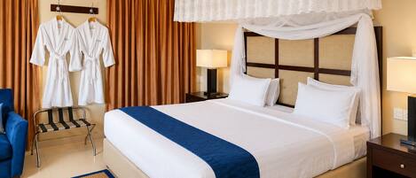 Habitación de hotel con una cama grande, un ventilador de techo, una mesita de noche con lámpara, una silla y una ventana con cortinas.