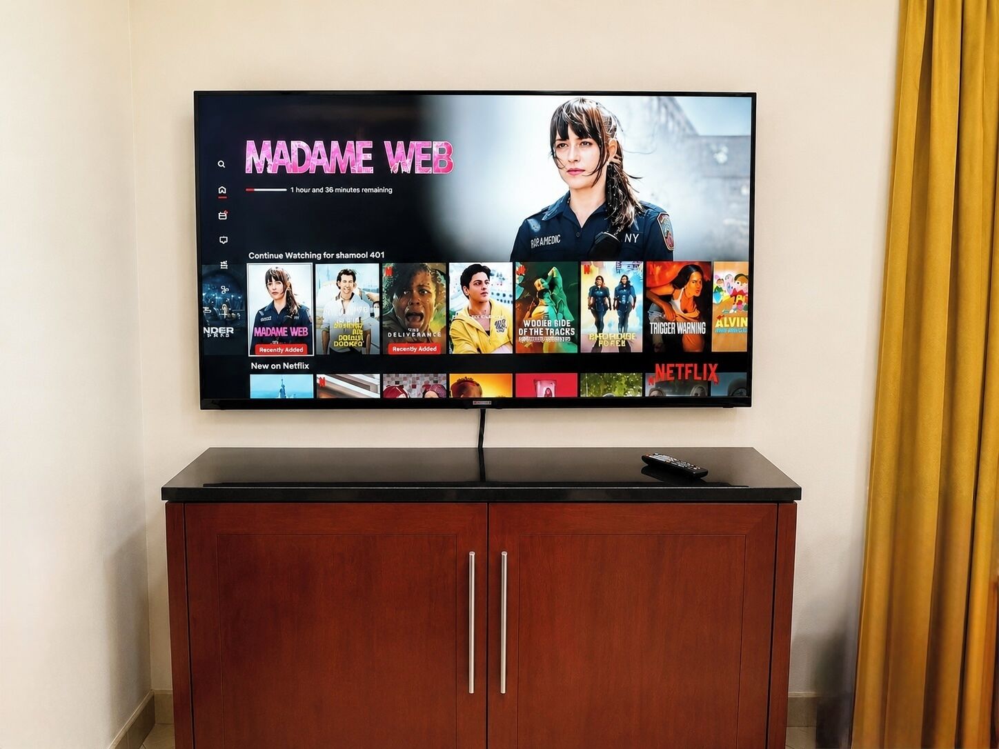 A flat-screen TV mounted on a wall above a wooden cabinet with a remote control on top.
