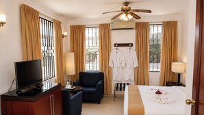 Habitación de hotel con cama, escritorio con televisor, silla, ventilador de techo y ventana con cortinas.