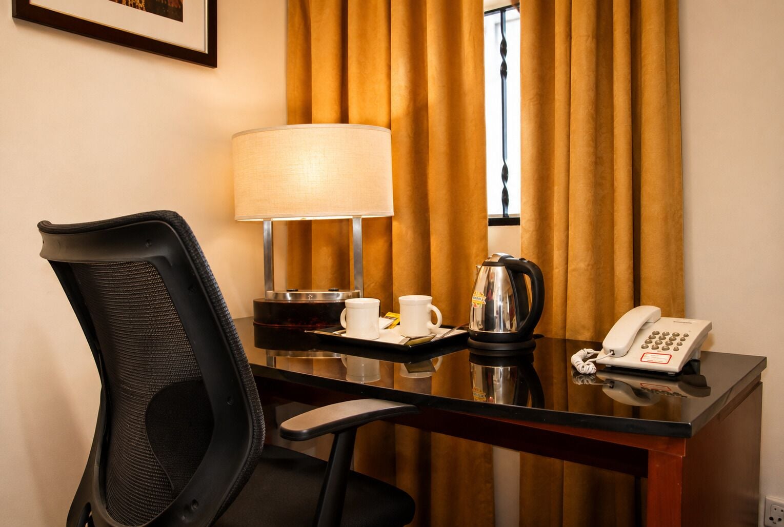 Habitació d'hotel amb escriptori, cadira, làmpada, cafetera, telèfon i finestra amb cortines.
