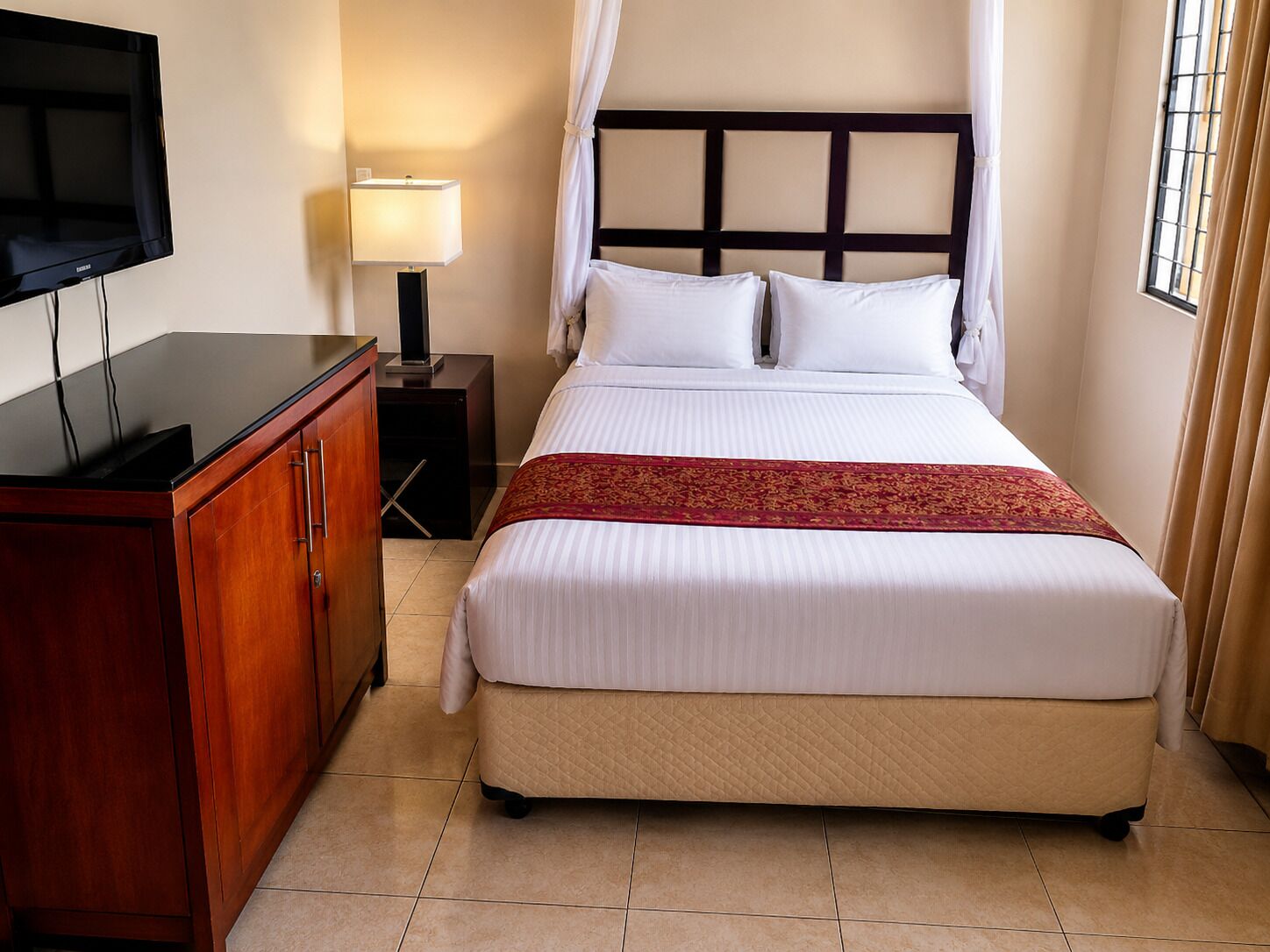 Une chambre d’hôtel avec un lit, une télévision, une table de chevet, une lampe et une fenêtre avec des rideaux.