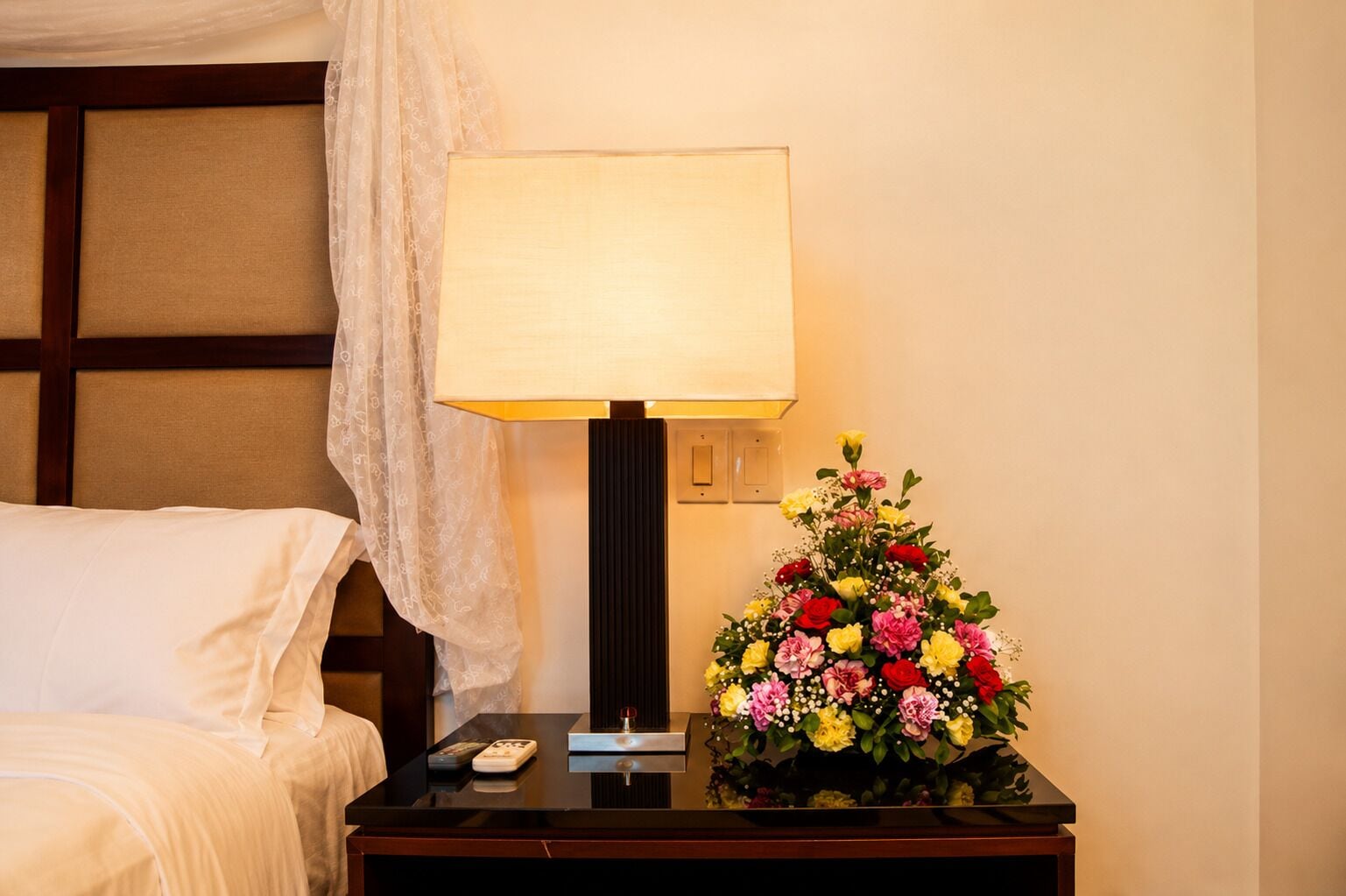 A hotel room with a bed, a nightstand with a lamp, and a floral arrangement.