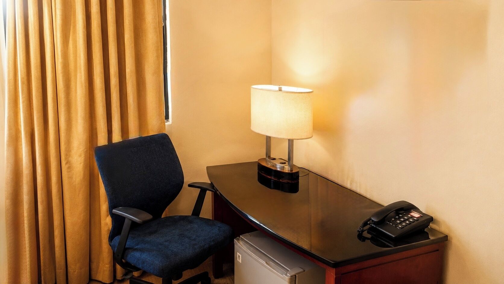 A hotel room with a desk, chair, lamp, phone, and a small refrigerator.