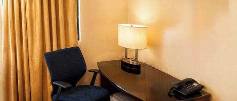 A hotel room with a desk, chair, lamp, phone, and a small refrigerator.