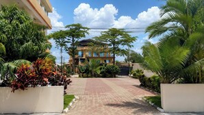 Un edificio residencial con entrada pavimentada, rodeado de exuberante vegetación y palmeras.