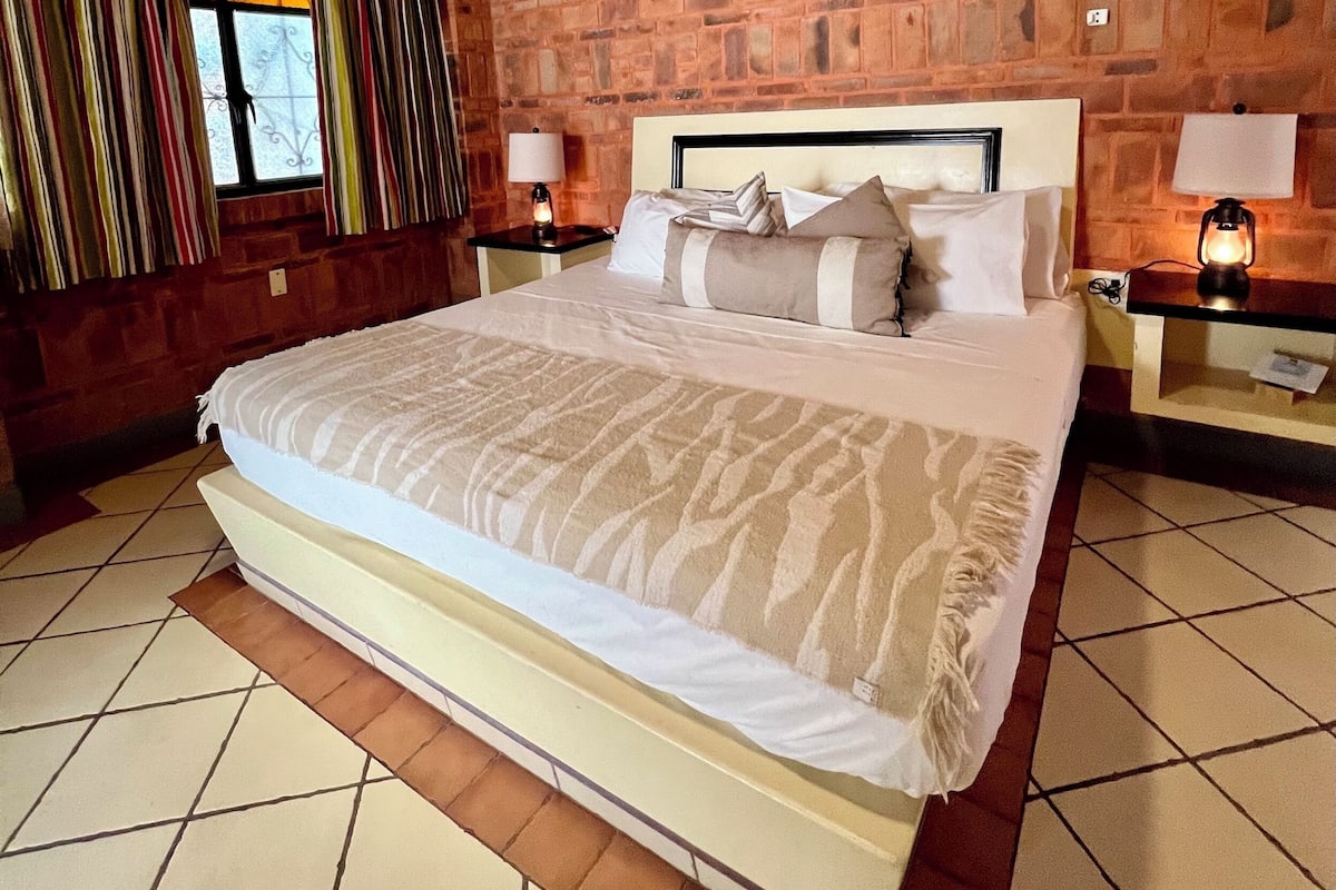 A hotel room with a brick wall, a large bed, two bedside tables with lamps, and a window with curtains.