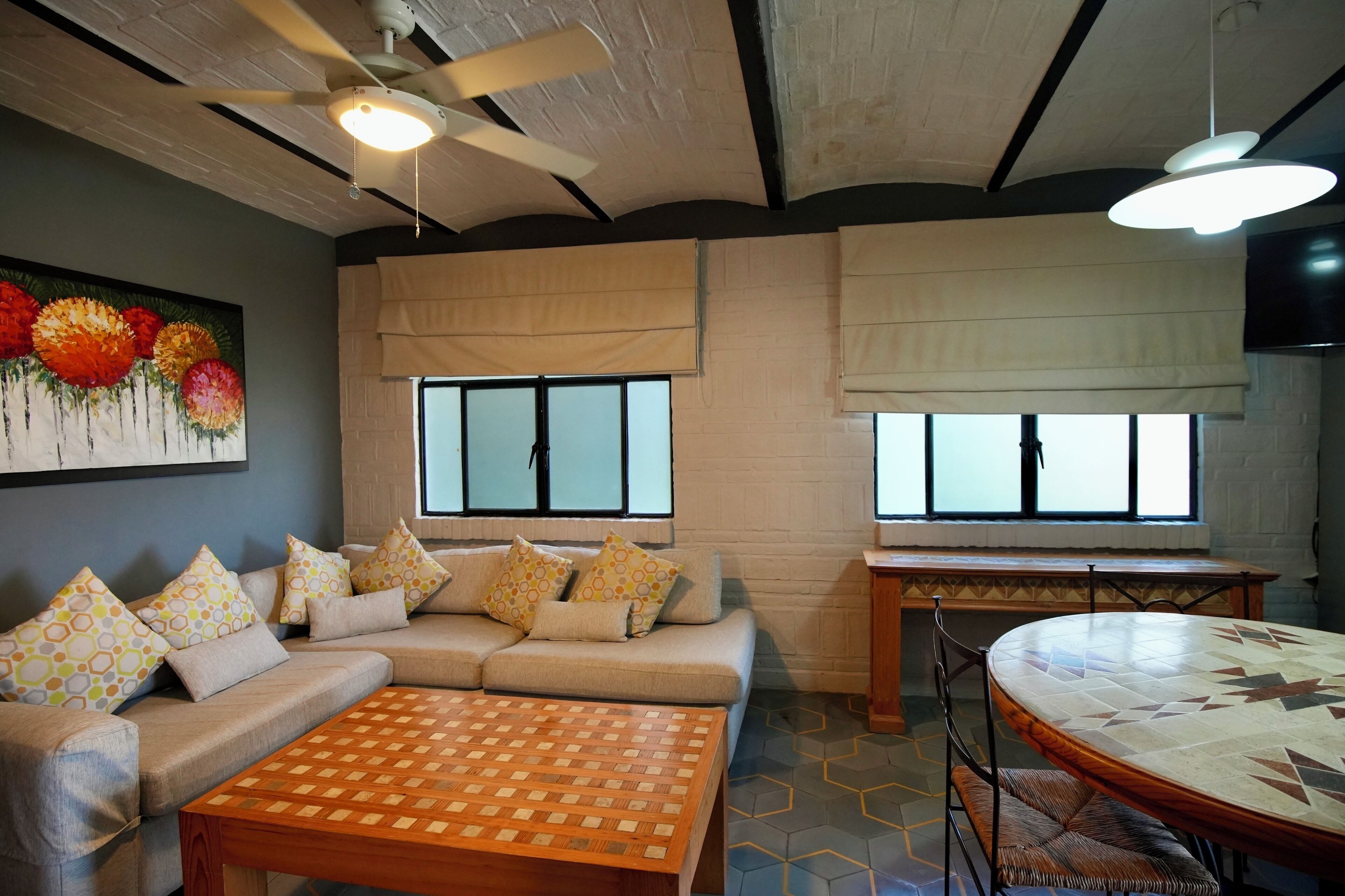 A living room with a sofa, coffee table, and dining table. There are windows with blinds, a ceiling fan, and a painting on the wall.