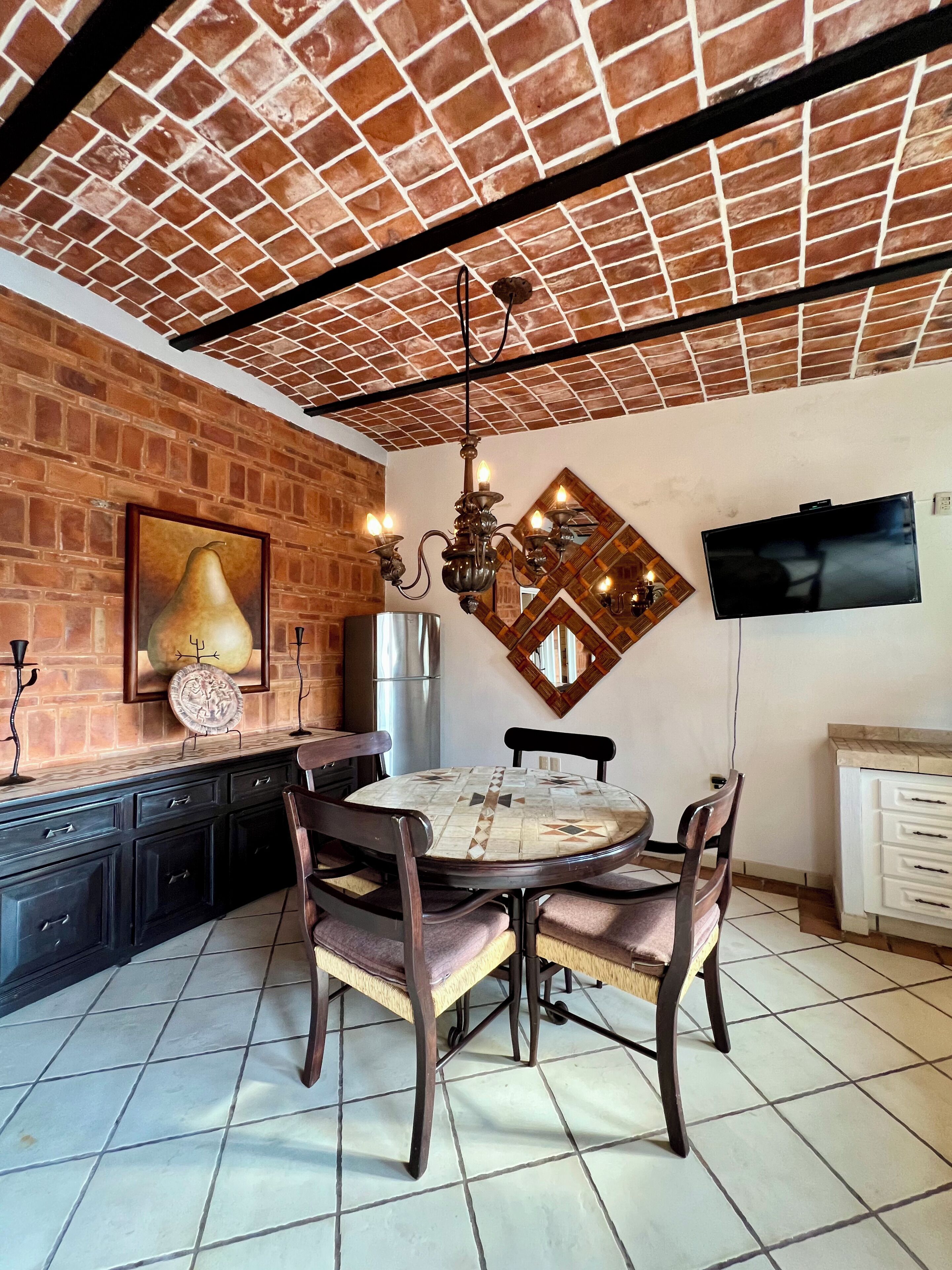 A dining area with a brick wall, a round table with chairs, a chandelier, and a television mounted on the wall.