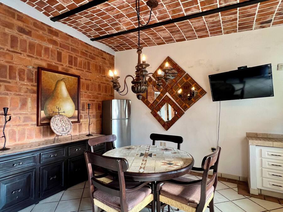 A dining area with a brick wall, a round table with chairs, a chandelier, and a television mounted on the wall.