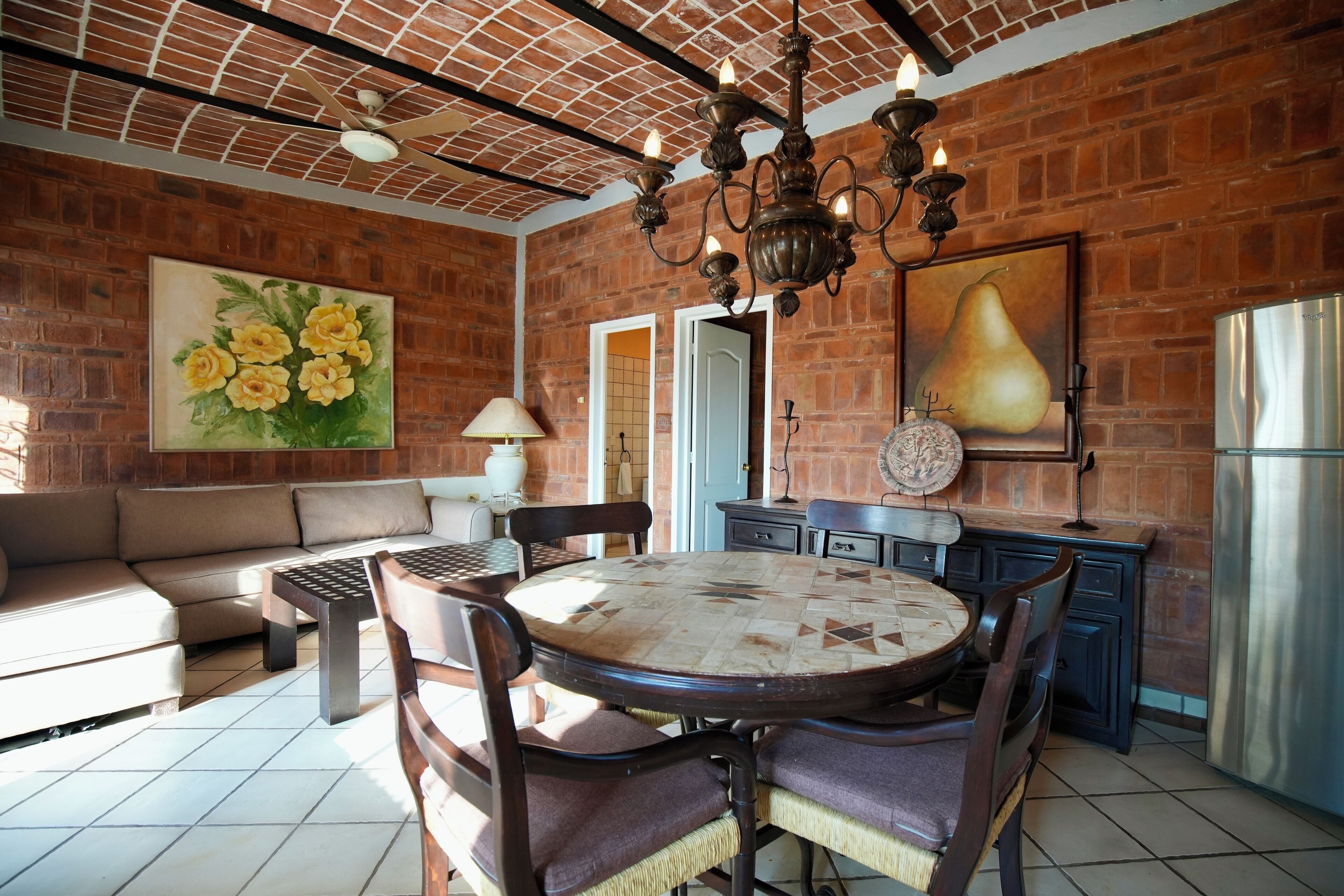 A dining area with a round table, chairs, a sofa, and a brick wall with a painting of flowers.