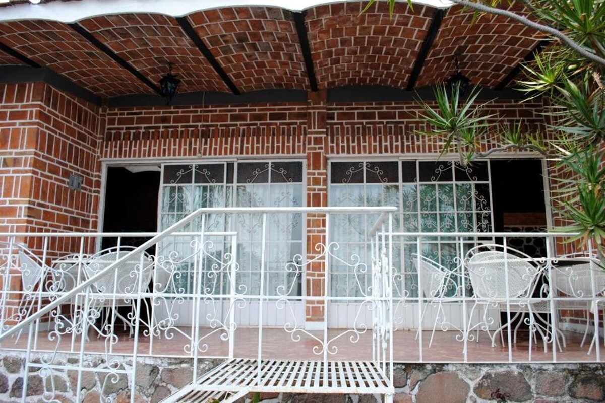 Een overdekt terras met een bakstenen muur, metalen hekwerk en witte metalen stoelen.
