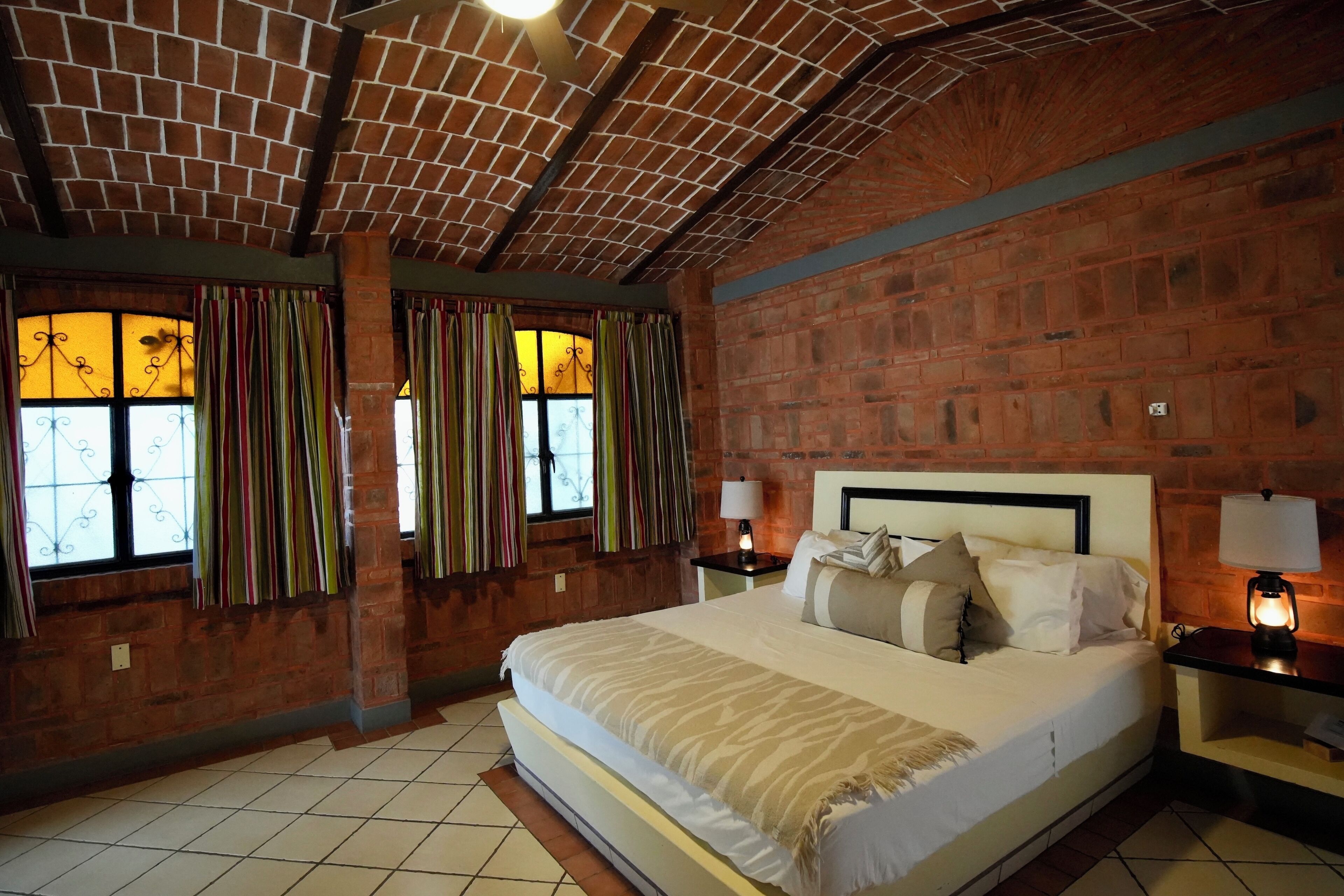 A hotel room with a brick wall, a large bed with multiple pillows, two bedside tables with lamps, and a window with curtains.