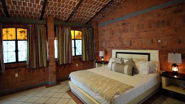 Une chambre d’hôtel avec un mur en briques, un grand lit avec de nombreux oreillers, deux tables de chevet avec des lampes et une fenêtre avec des rideaux.