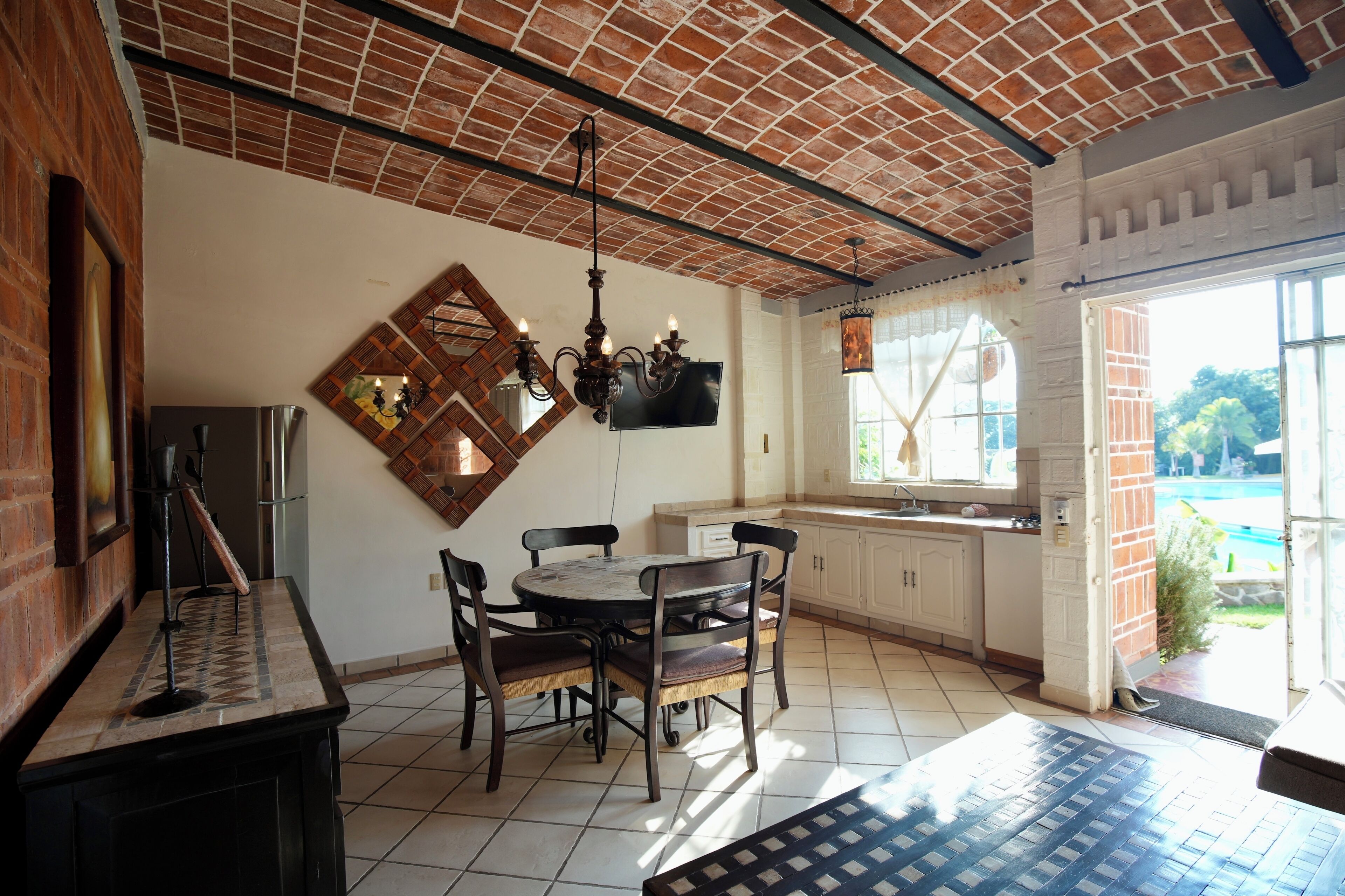 Una cocina con techo de ladrillo, armarios blancos, una mesa de comedor con sillas y un televisor en la pared.