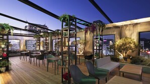 An outdoor seating area with wooden flooring, green chairs, and tables, surrounded by plants and a city view at dusk. - Moxy Seoul Insadong (Seoul)